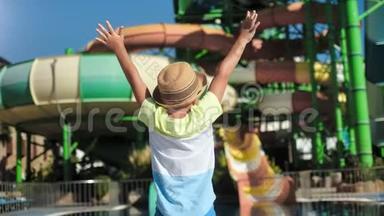 晒太阳的小男孩戴着帽子跳着，举起手在户外水上乐园里欢度节日